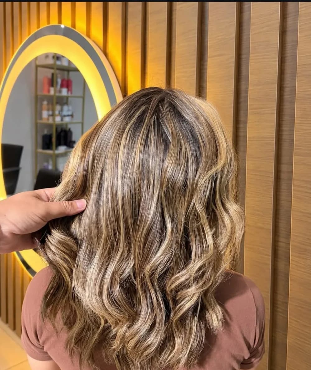 Cabelo com mechas iluminadas e ondas, fotografado de costas diante do espelho iluminado do salão